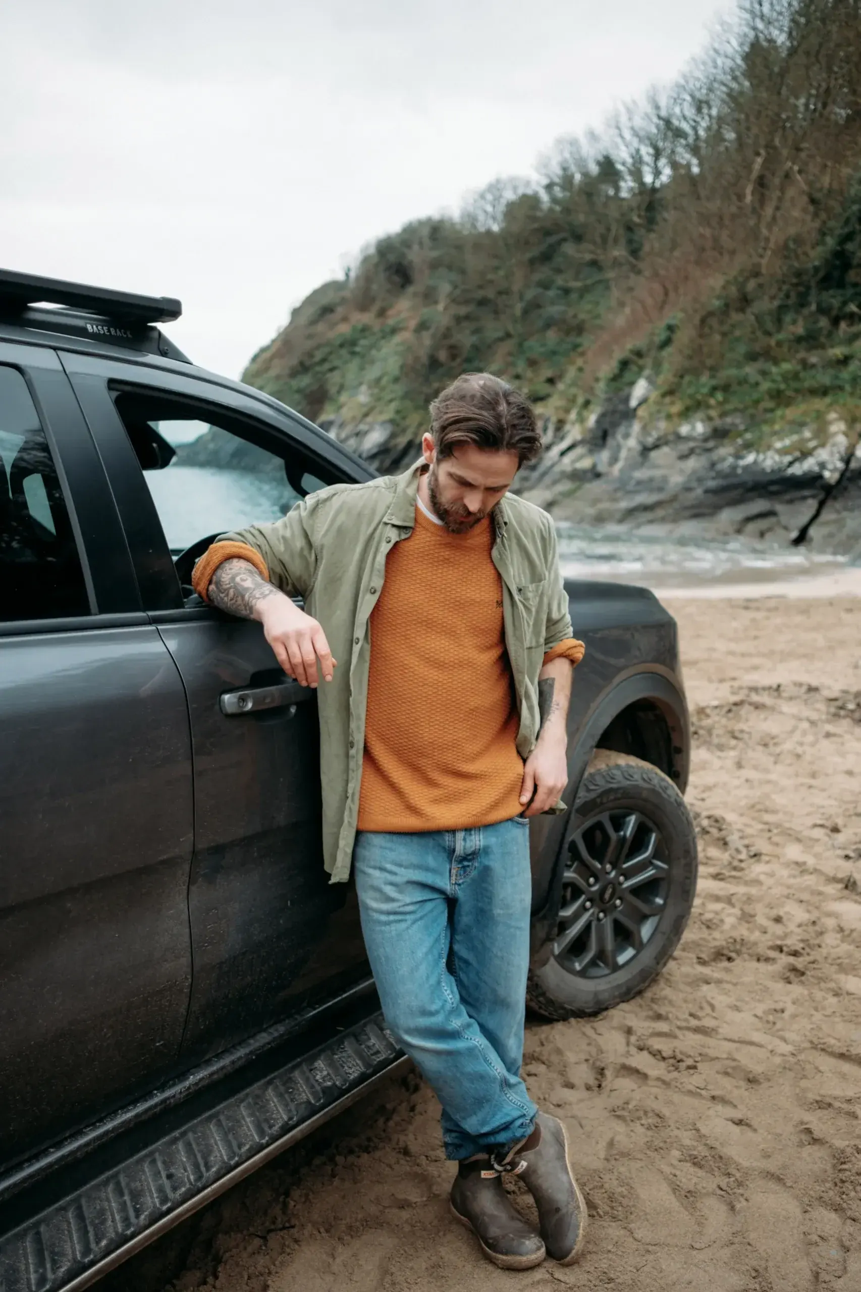 James Strawbridge leaning against a Ford Ranger Wildtrak with ARB base rack on a sandy Cornish beach cove