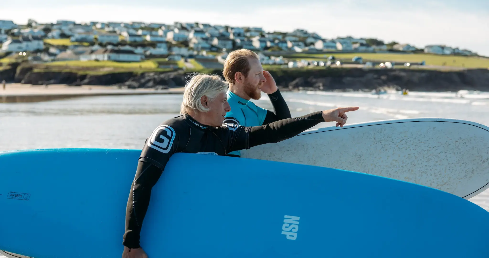George and a surfer carrying blue surfboards across Polzeath beach during a Perfect Stays concierge film
