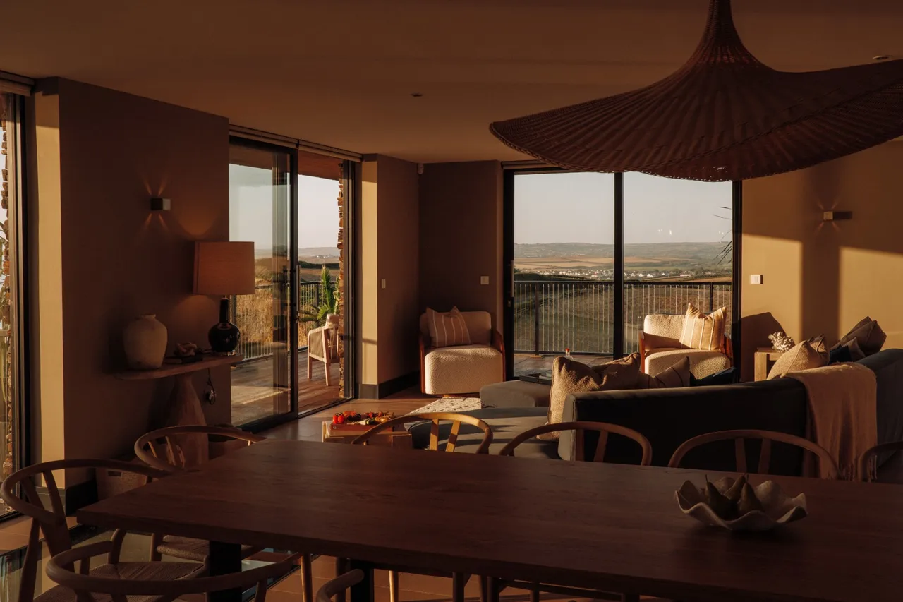 Open-plan living and dining room at Namuri in low golden light, with glass doors onto the balcony and the North Cornwall countryside beyond