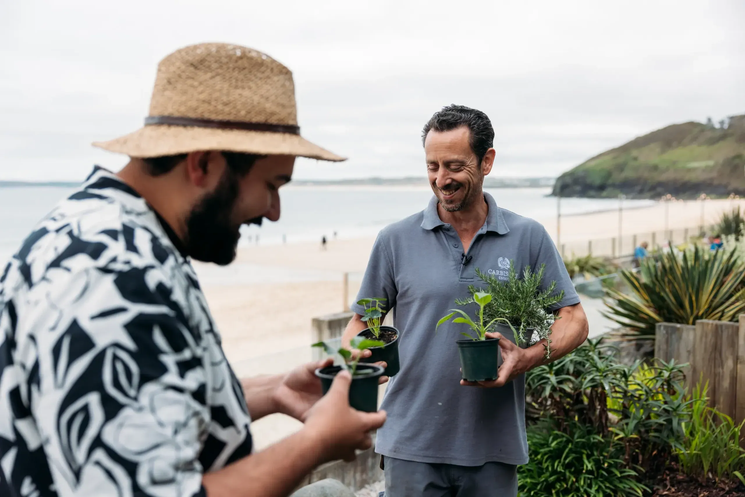 Chris the head gardener being interviewed by Joe from Joe’s Garden TikTok channel at Carbis Bay Estate, holding fresh herb plants with the beach and Cornish coastline behind them