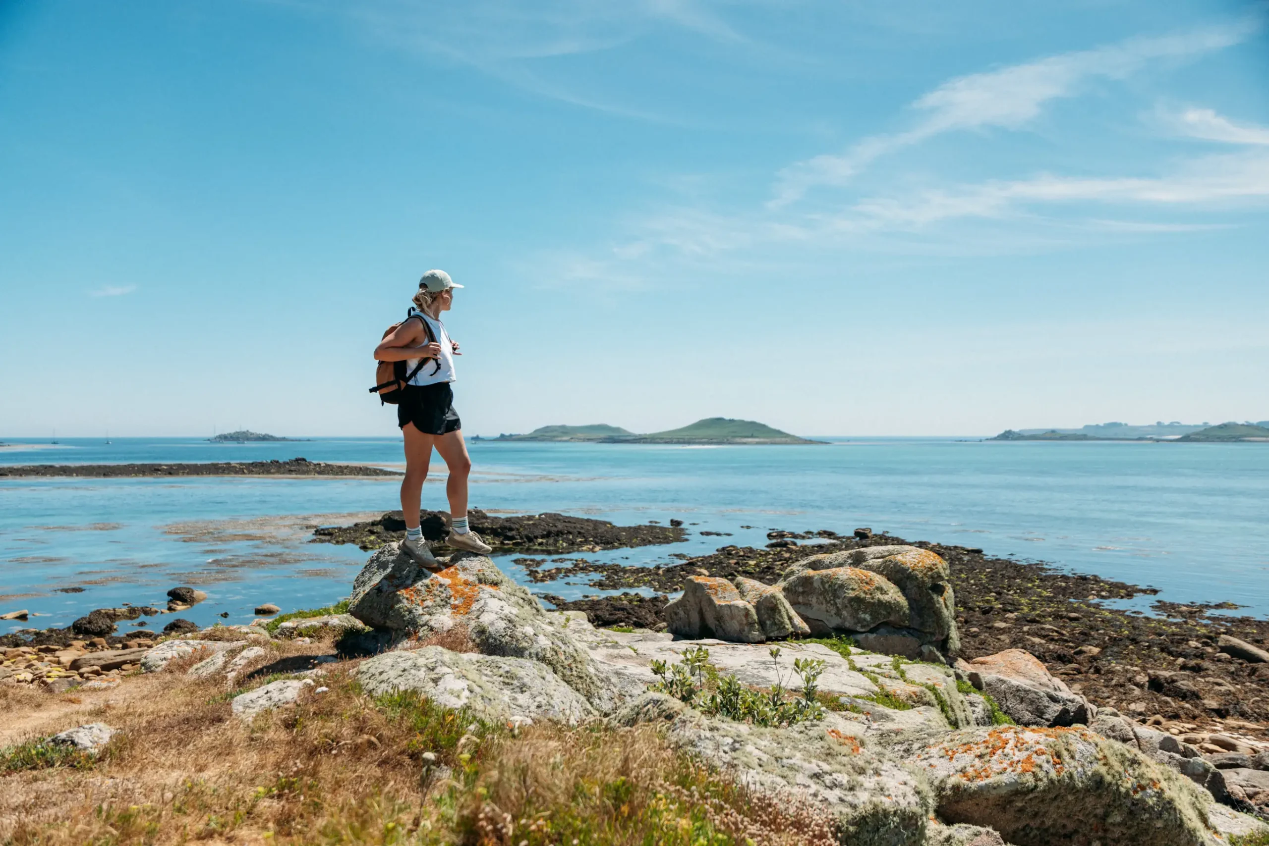 Woman standing on rocky shoreline of St Martin’s, Isles of Scilly, looking out over turquoise waters on a clear summer day – PixelRain destination film production.