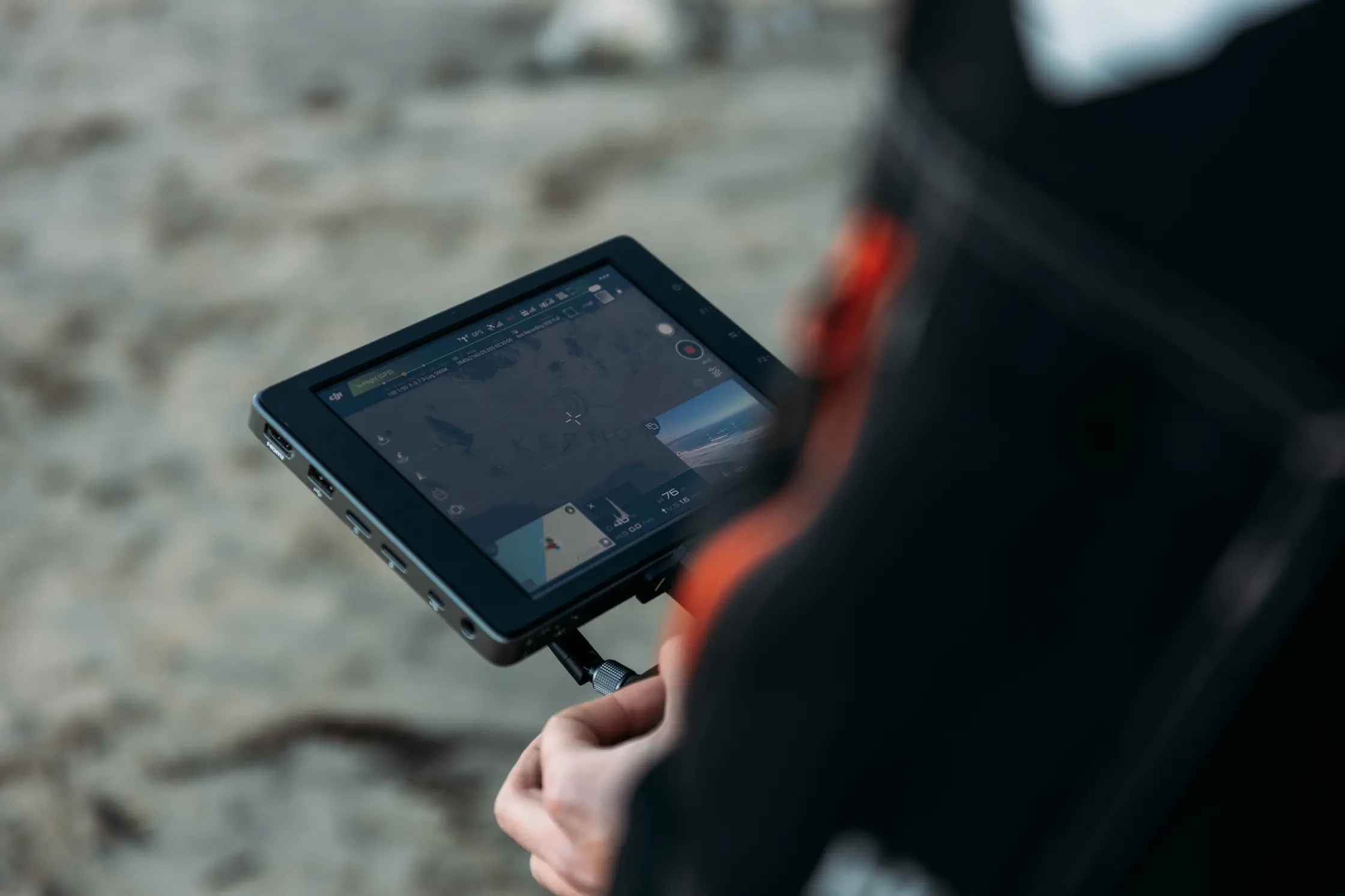 Drone operator monitoring the live feed on a DJI controller screen during aerial filming of the Kernow Asset Management sand art shoot on a Cornish beach, by PixelRain